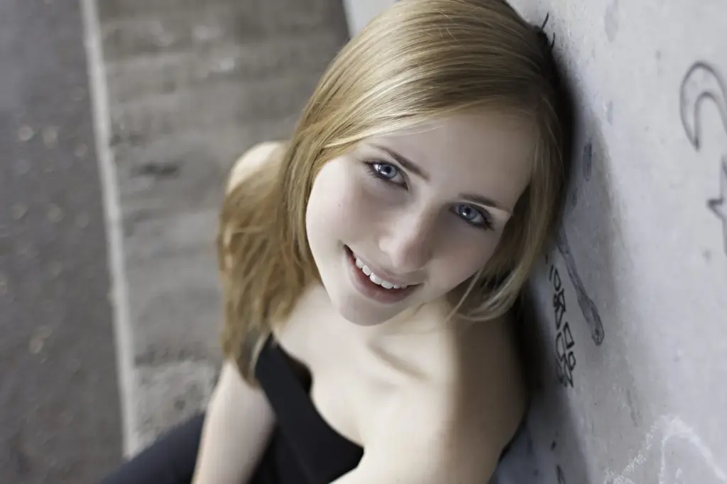 junge Frau mit langen blonden Haaren Hand in urbanen Umgebung sitzend hochschauend Portr&auml;t - color