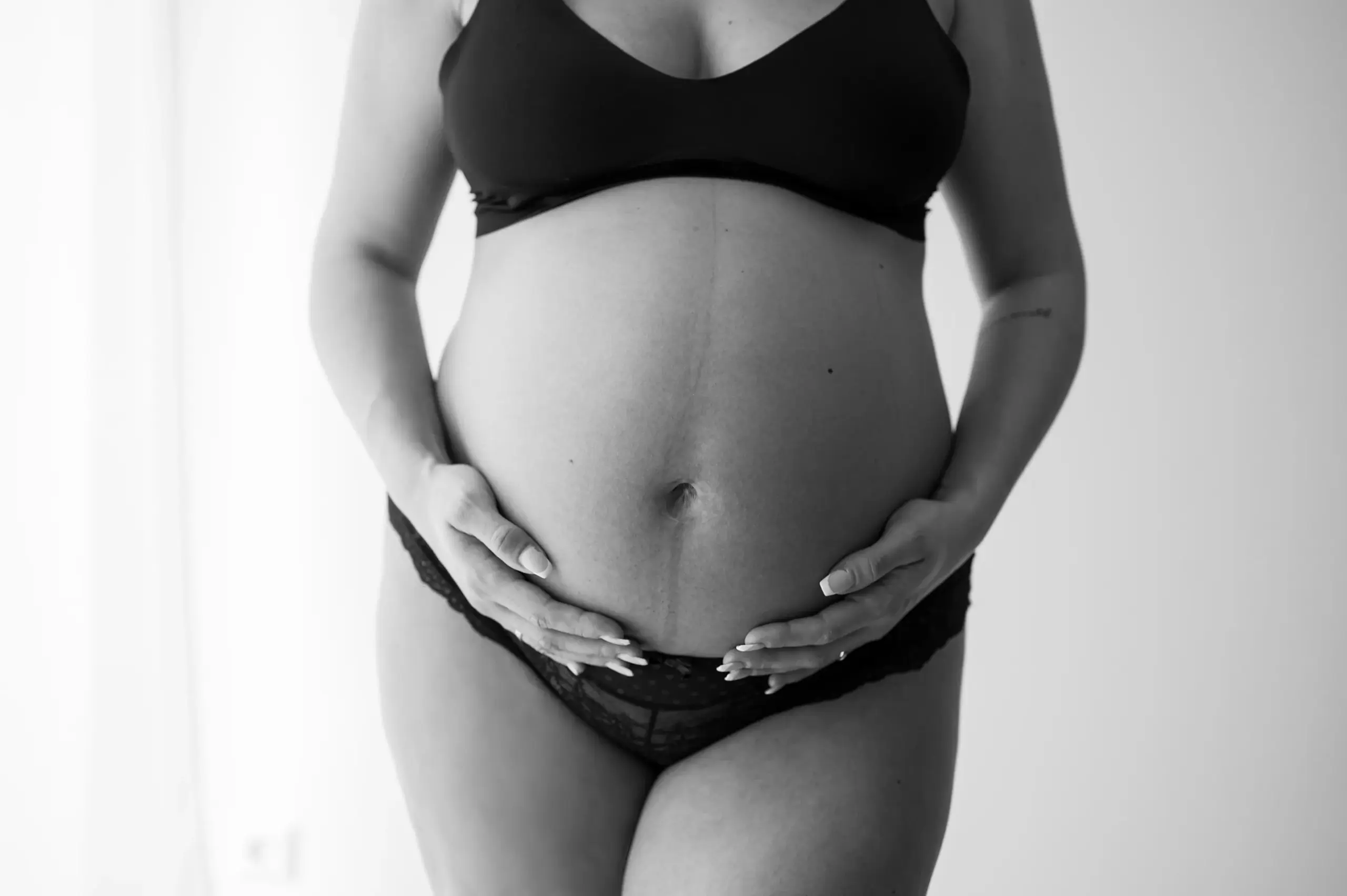 Detailaufnahme Bauch schwanger in schwarzer Spitzenhose und Bustier Hände locker auf dem Bauch sanftes Licht luftig elegant sw